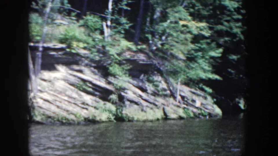 1964 Wisconsin home-movie frame with a moving view of trees, exposed rock, and lake water.