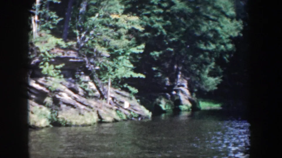 1964 Wisconsin 8mm frame showing dense woods, layered shoreline rock, and calm water.