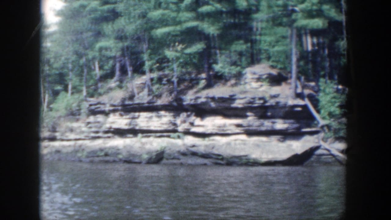 Wooded rock shoreline beside calm river water with dark 8mm frame borders.
