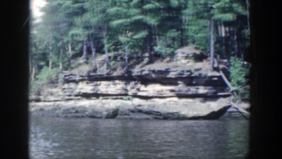 1964 Wisconsin 8mm frame showing layered rock banks, dense trees, calm river water, and dark film-frame edges.