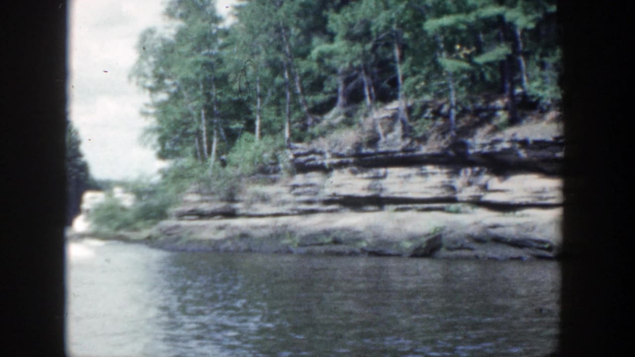 1964 Wisconsin river view with wooded rock ledges on both banks and a pale opening down the waterway.