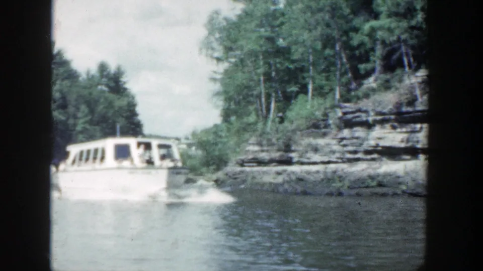 1964 Wisconsin 8mm frame with a white tour boat carrying passengers past rock cliffs and wooded shoreline.