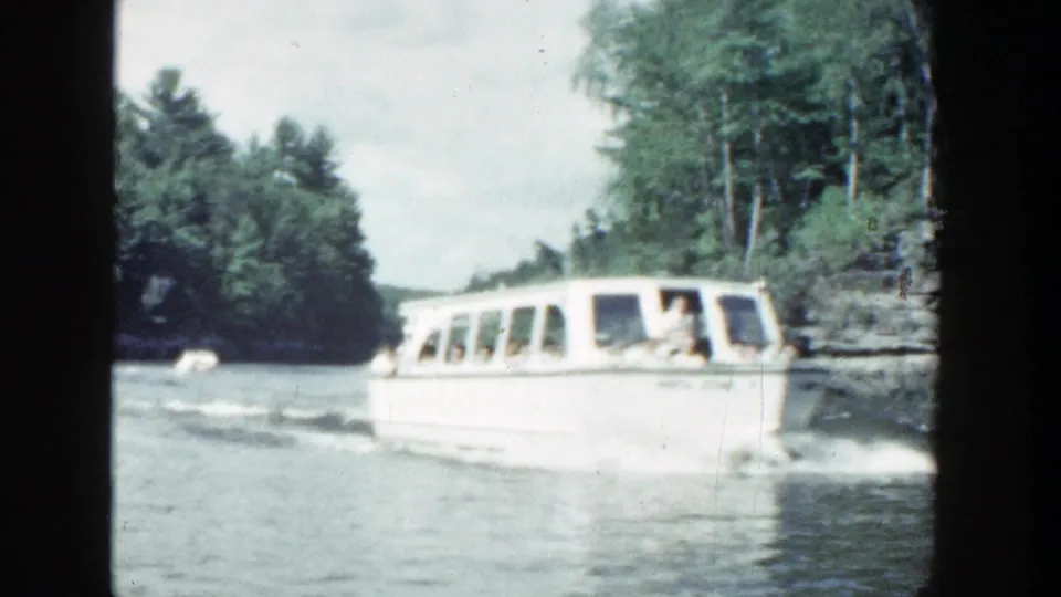 1964 Wisconsin home-movie frame showing a white passenger boat, visible riders, wake, trees, and layered rock along the river.