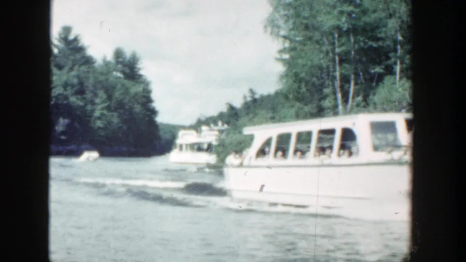 1964 Wisconsin 8mm frame showing a white sightseeing boat moving past trees on a river, with passengers visible inside.