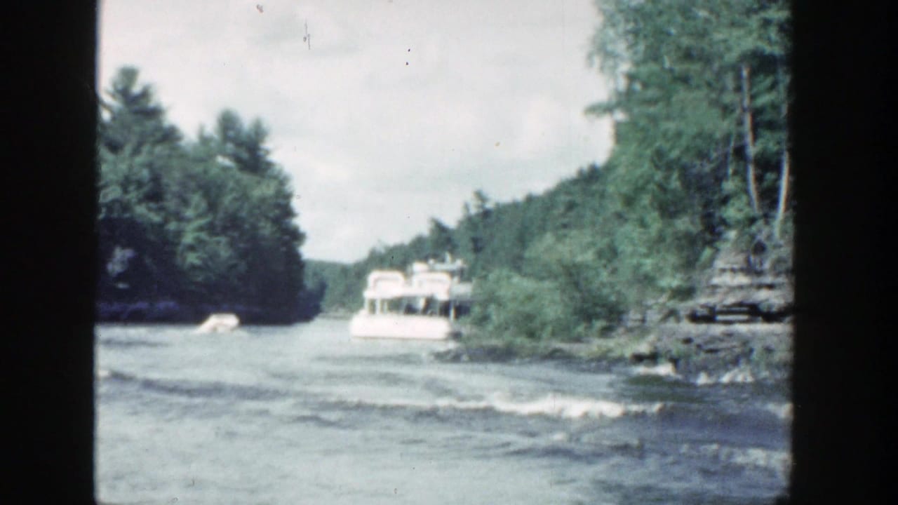 A tour boat recedes along a tree-lined river beside a rocky shoreline.