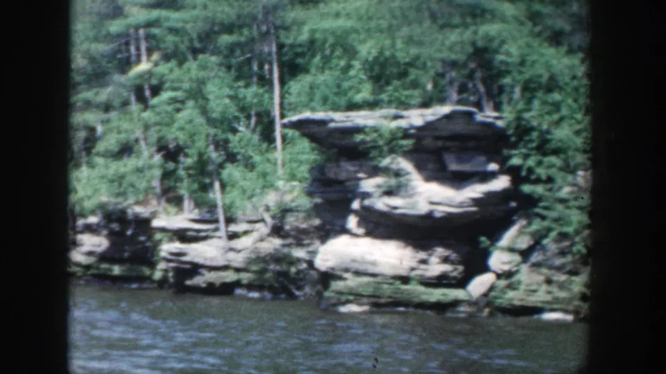 1964 Wisconsin 8mm frame showing a rocky wooded shoreline and calm river water from a moving boat perspective.