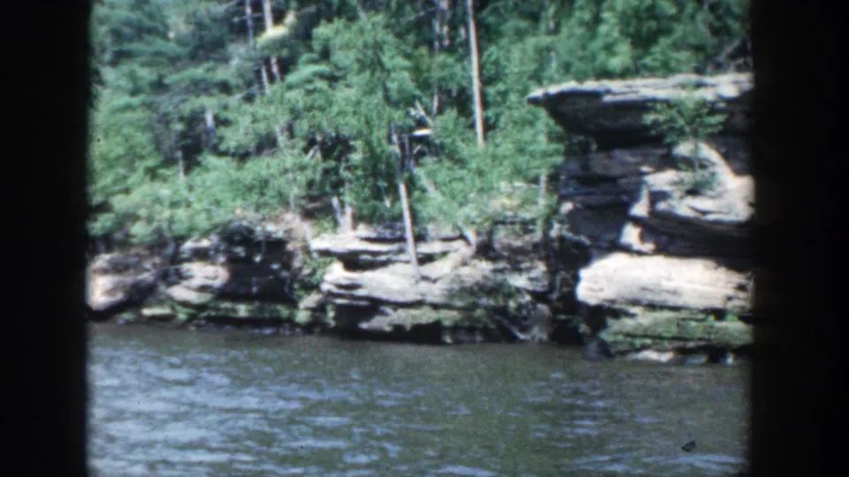 1964 Wisconsin riverside scenery with water, trees, and pale rock layers along the bank, filmed in a home-movie style.