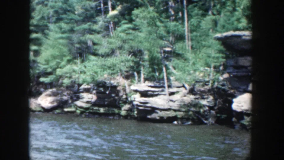 1964 Wisconsin 8mm frame of a wooded rocky shoreline seen from across rippling river water.