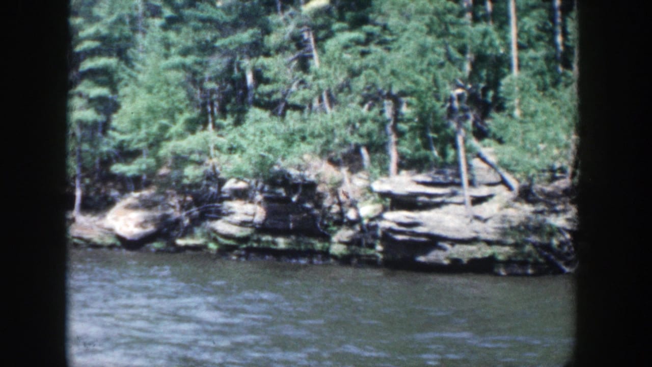 1964 Wisconsin 8mm view of layered shoreline rocks and dense trees from a moving river perspective.