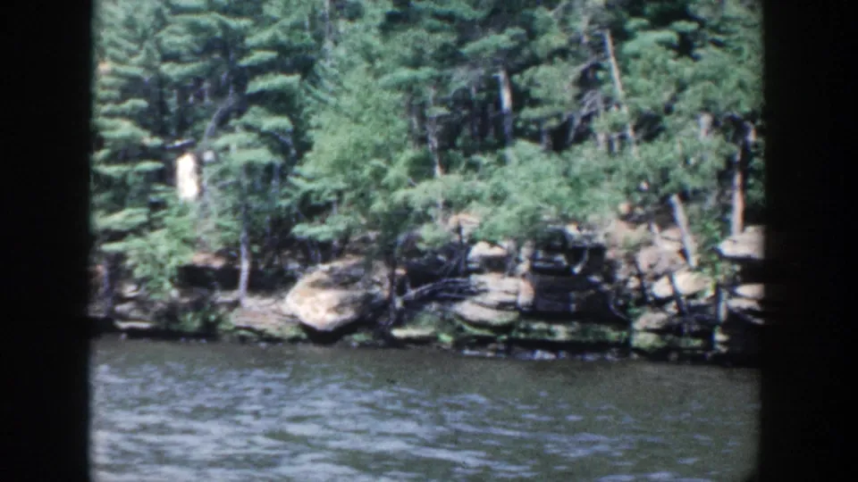 1964 Wisconsin home-movie frame showing a rocky wooded bank beside dark rippling river water.