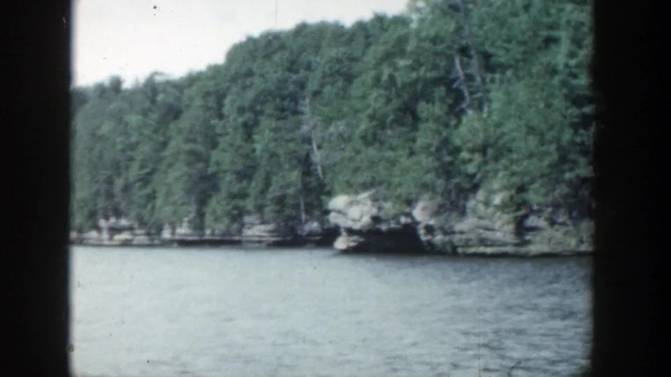 1964 Wisconsin 8mm frame with a wider river channel, forested shore, rocky outcrop, and pale sky.
