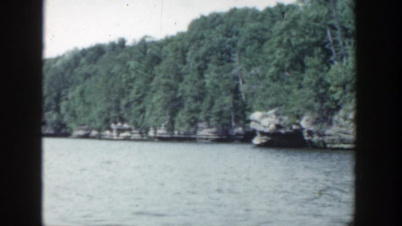 1964 Wisconsin home-movie frame showing calm blue-gray lake water, dense green trees, and rocky shoreline seen through dark film-frame edges.