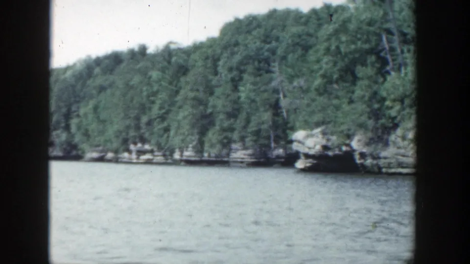 1964 Wisconsin home-movie frame showing calm blue-gray lake water, dense green trees, and rocky shoreline seen through dark film-frame edges.