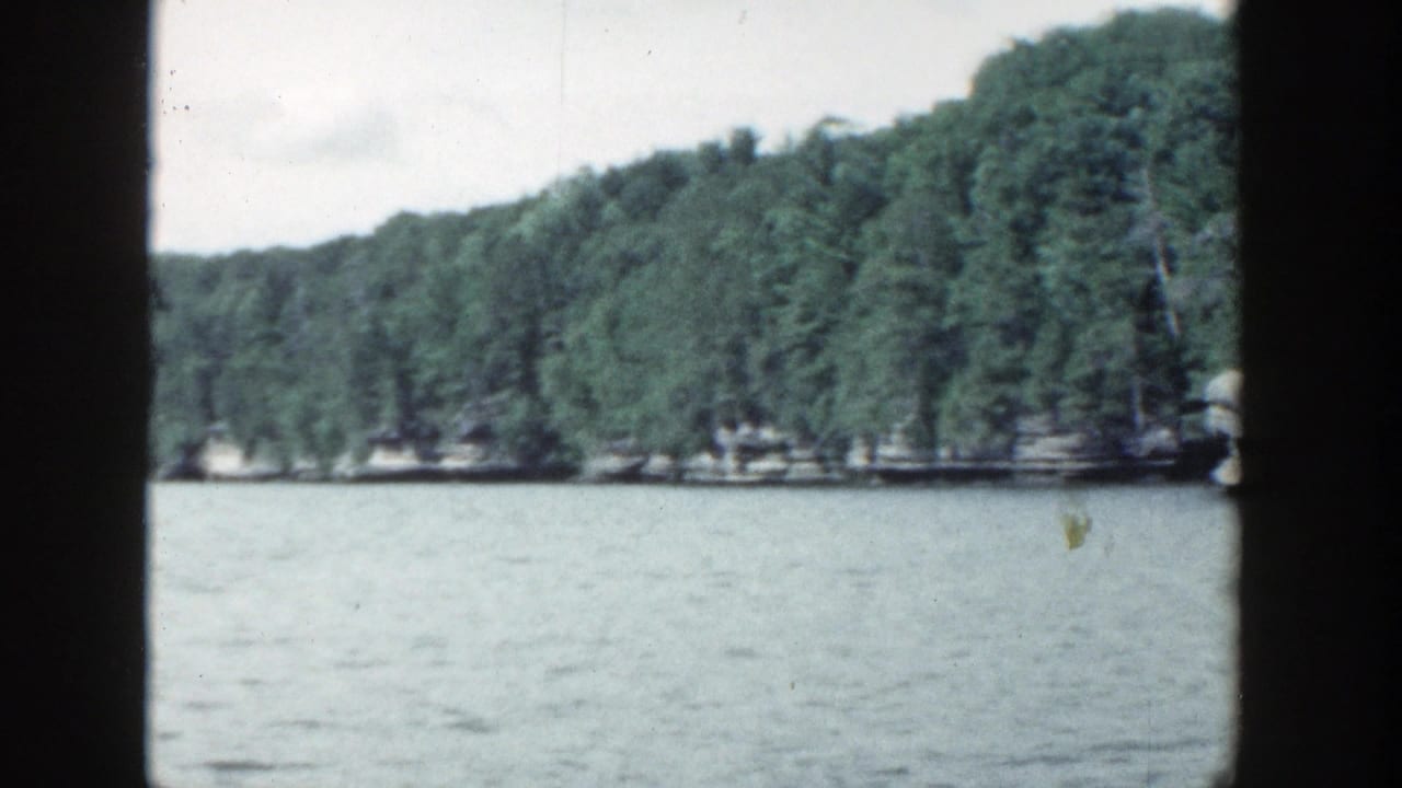 1964 Wisconsin lake view with rippled water, dense trees along the far shore, pale sky, and a dark vertical obstruction near the center of the frame.