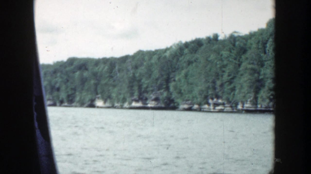 1964 Wisconsin home-movie frame showing a broad lake surface, low wooded shoreline, pale sky, and dark edge borders from the 8mm scan.