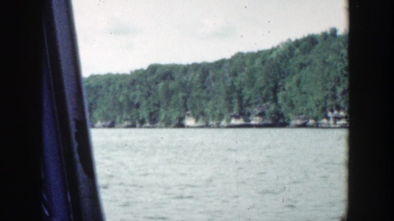 1964 Wisconsin lakeshore footage with calm water in the foreground, a distant green tree line, pale sky, and a dark vertical band crossing the frame.