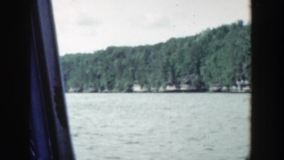 1964 Wisconsin lakeshore footage with calm water in the foreground, a distant green tree line, pale sky, and a dark vertical band crossing the frame.