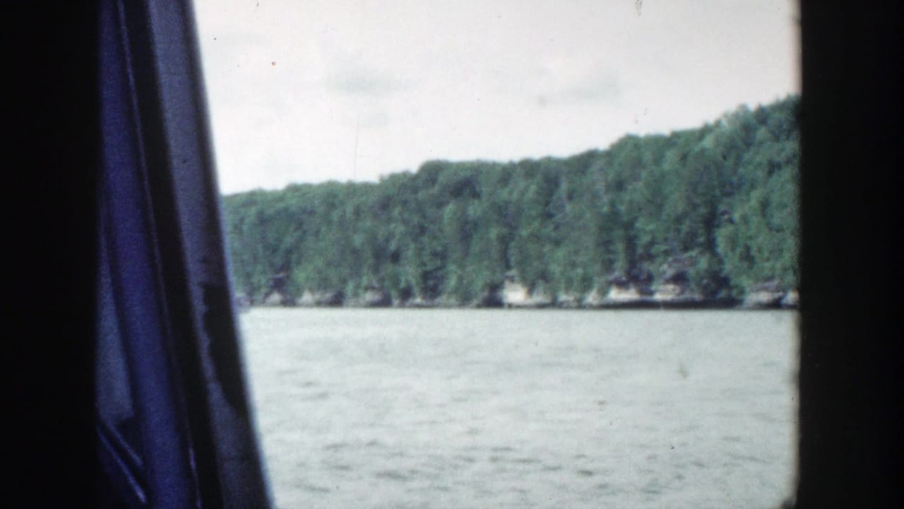 Lake and wooded shoreline seen through a dark vehicle window with a blue curtain.