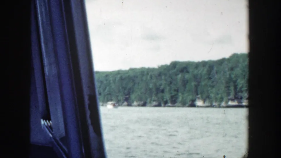 Wisconsin lakeshore with dense trees and calm water, seen from inside a moving vehicle in 1964 home-movie footage.