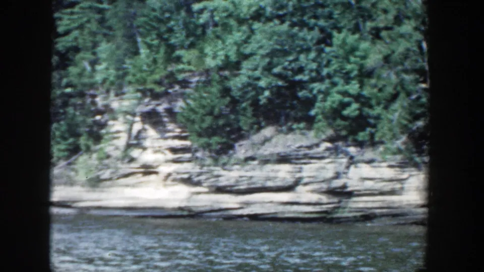 1964 Wisconsin shoreline detail showing layered rock ledges, dense trees, and lake water from a moving passenger view.