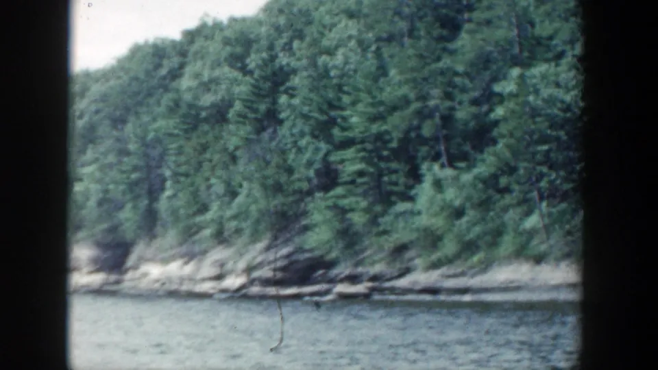 1964 Wisconsin 8mm frame with motion-softened trees, rocky bank, and river water from a moving boat perspective.