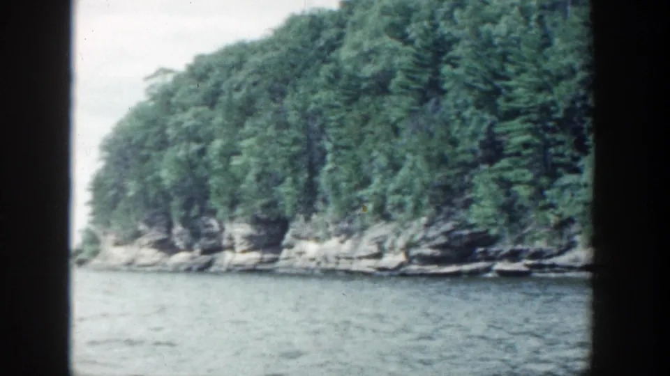 1964 Wisconsin river view showing open water, a curving wooded shoreline, and exposed rock ledges.