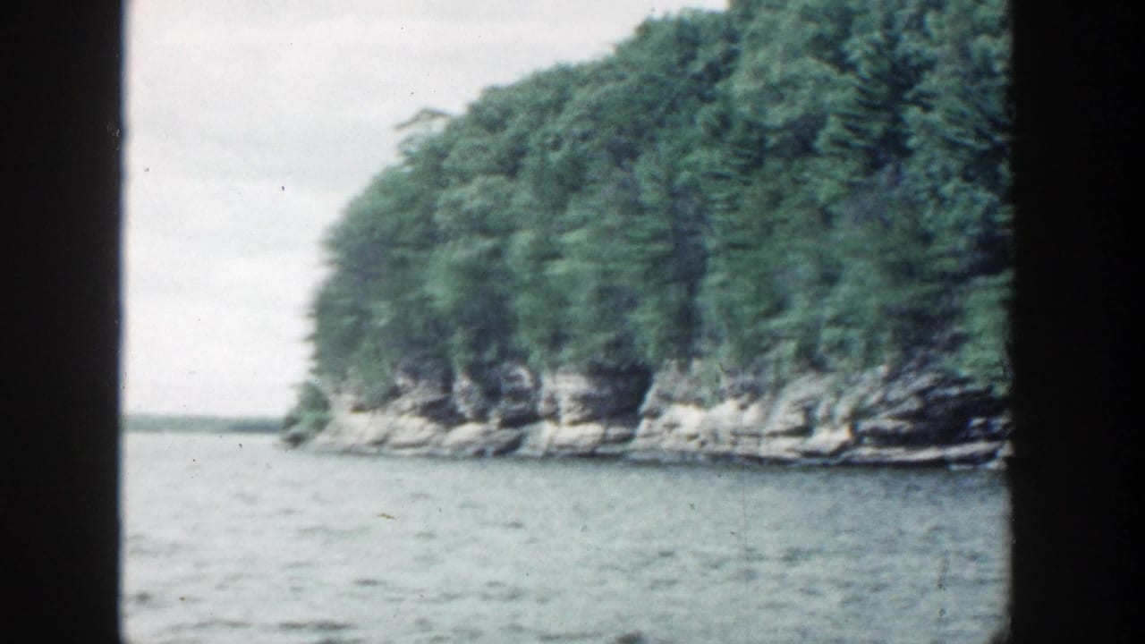 1964 Wisconsin 8mm frame showing a tree-covered rocky shoreline viewed from calm water beneath a pale overcast sky.
