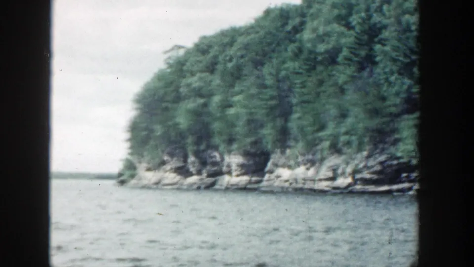 1964 Wisconsin 8mm frame showing a tree-covered rocky shoreline viewed from calm water beneath a pale overcast sky.
