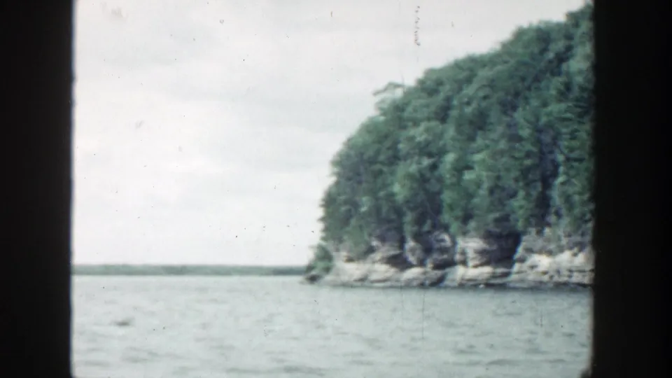 1964 Wisconsin 8mm view of a forested rocky point bordering open water, with the contact-sheet frame edge visible.