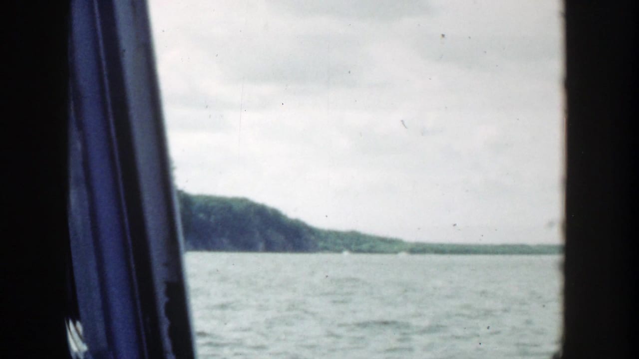 1964 Wisconsin 8mm boat view looking across calm lake water toward a distant tree-lined shore, with dark window framing and blue fabric at left.