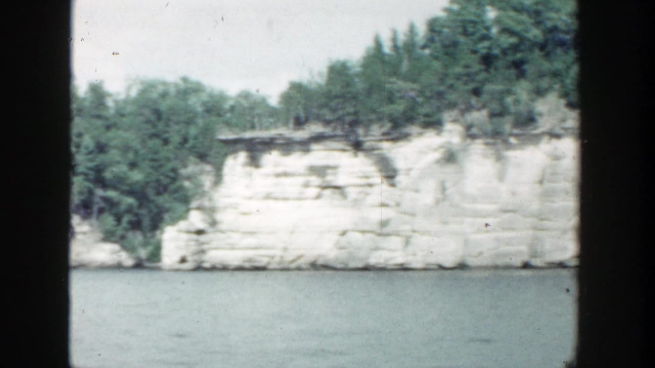 Pale cliff face and trees along a lake shoreline seen through dark boat-window framing.
