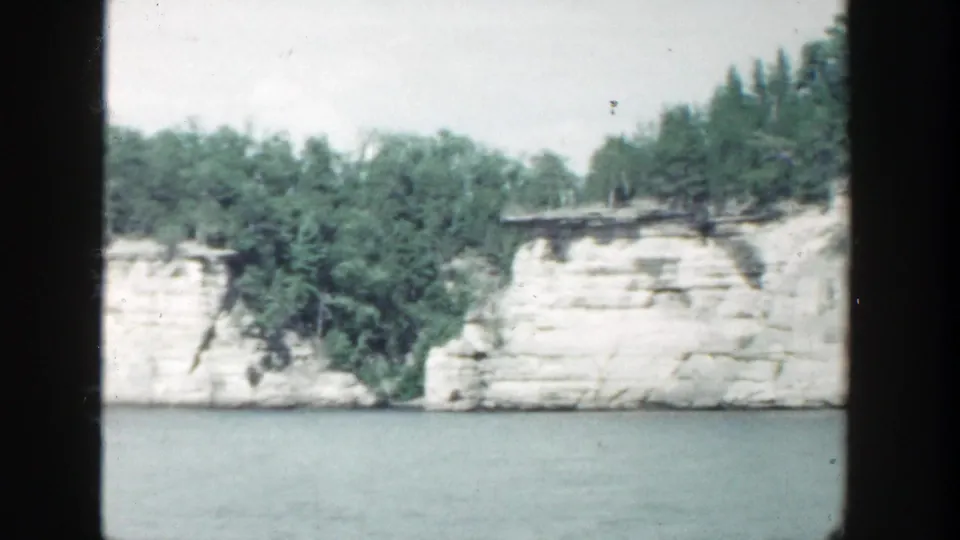 1964 Wisconsin 8mm footage frame showing white limestone cliffs, trees, and calm lake water from a passenger's boat-window viewpoint.