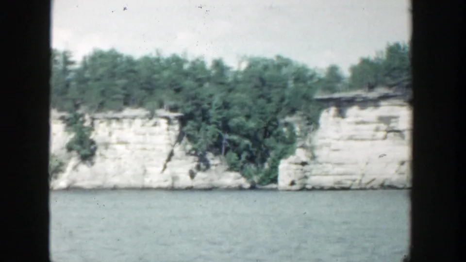 1964 Wisconsin lake scene with pale shoreline cliffs, dense trees, and calm water, captured in soft color 8mm from a boat.