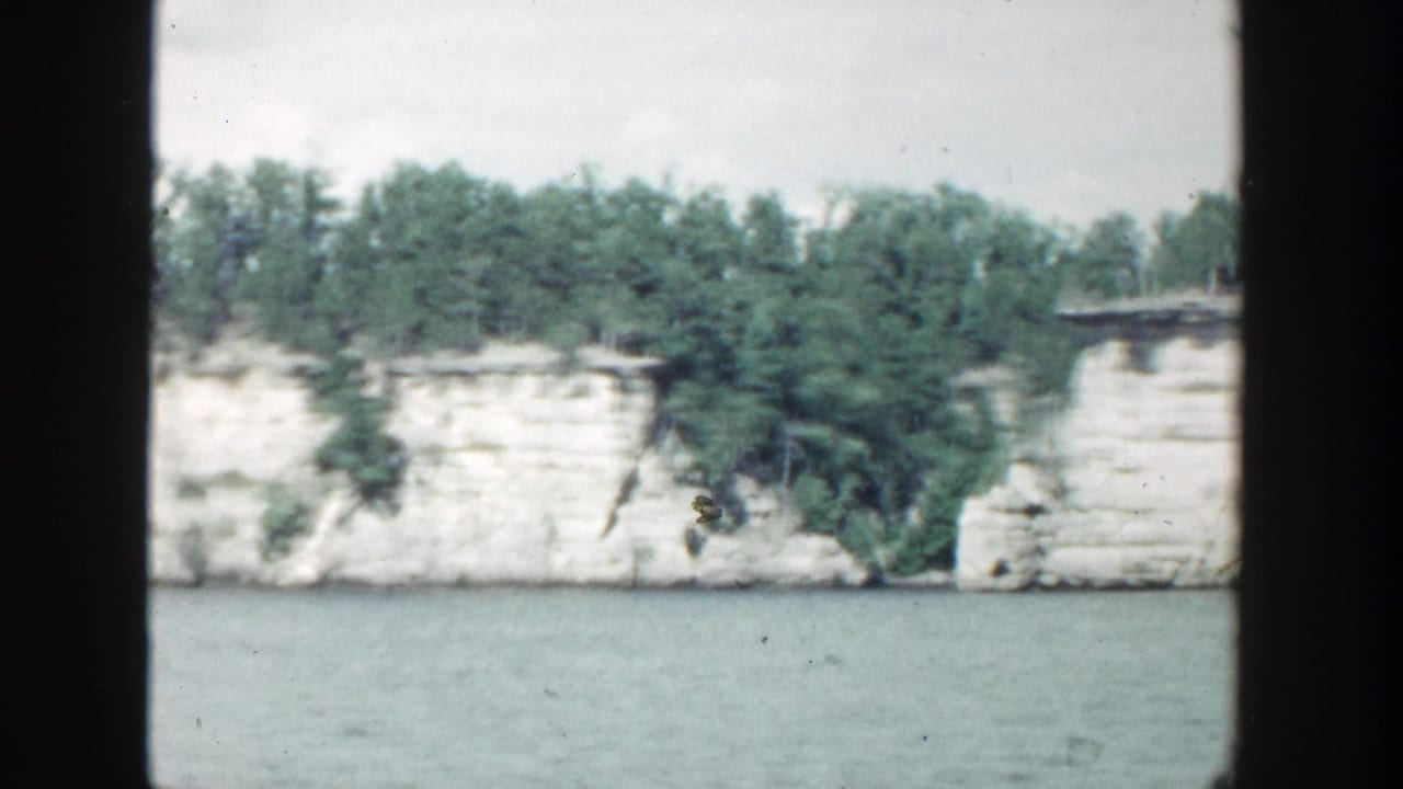 Pale stone cliff with trees above and calm water below, framed by dark film borders.