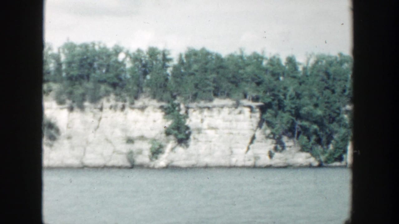 White rock face and tree-covered shoreline seen across calm water.