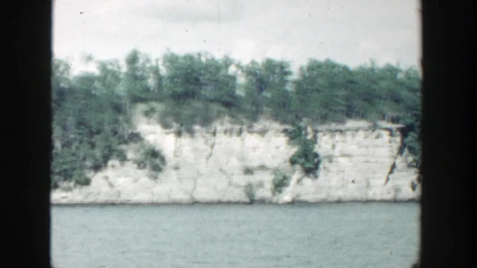 1964 Wisconsin 8mm frame of a layered pale cliff with dense greenery above and blue-green water in the foreground.