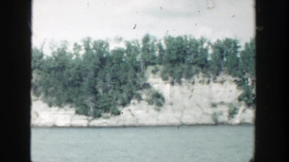 1964 Wisconsin home-movie view of a tree-lined white stone wall and calm water, with soft grain and dark border edges.