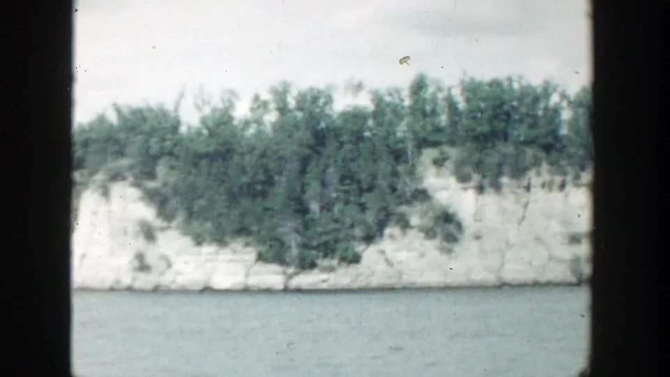 1964 Wisconsin 8mm frame showing a pale rocky lakeshore with dense green trees above calm water.