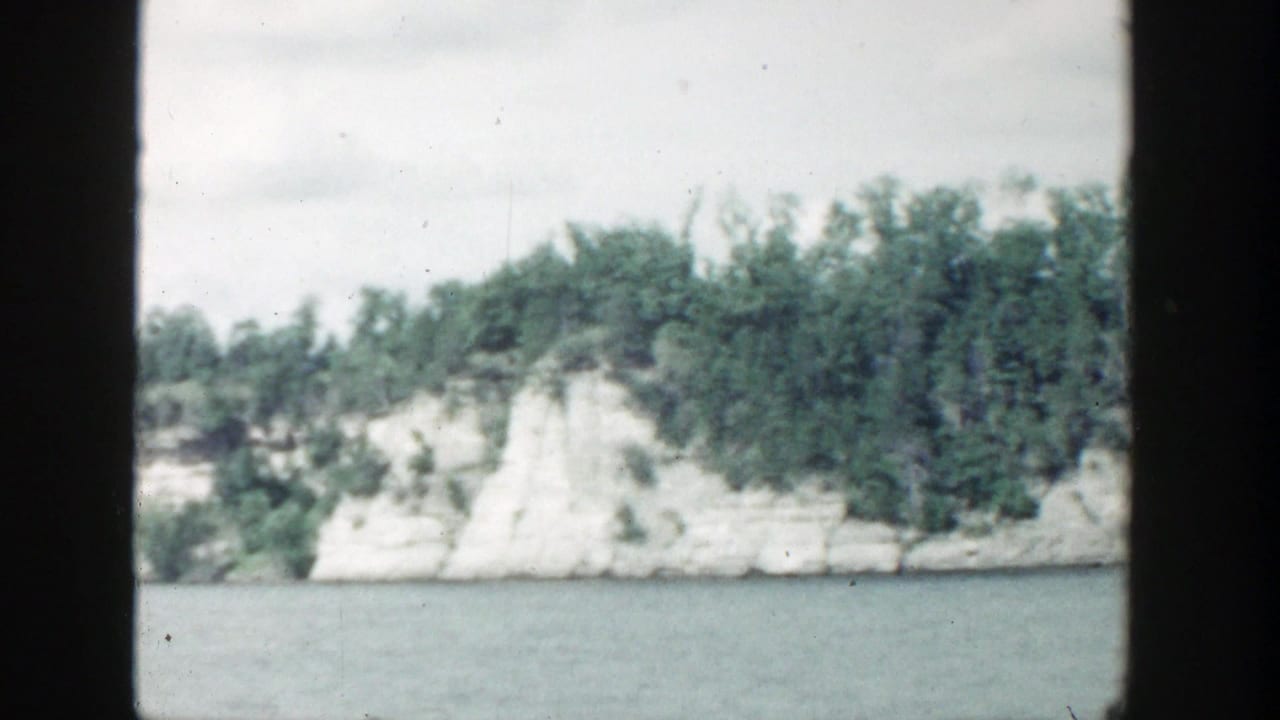 Tree-covered pale rock shoreline viewed across water with dark film borders.