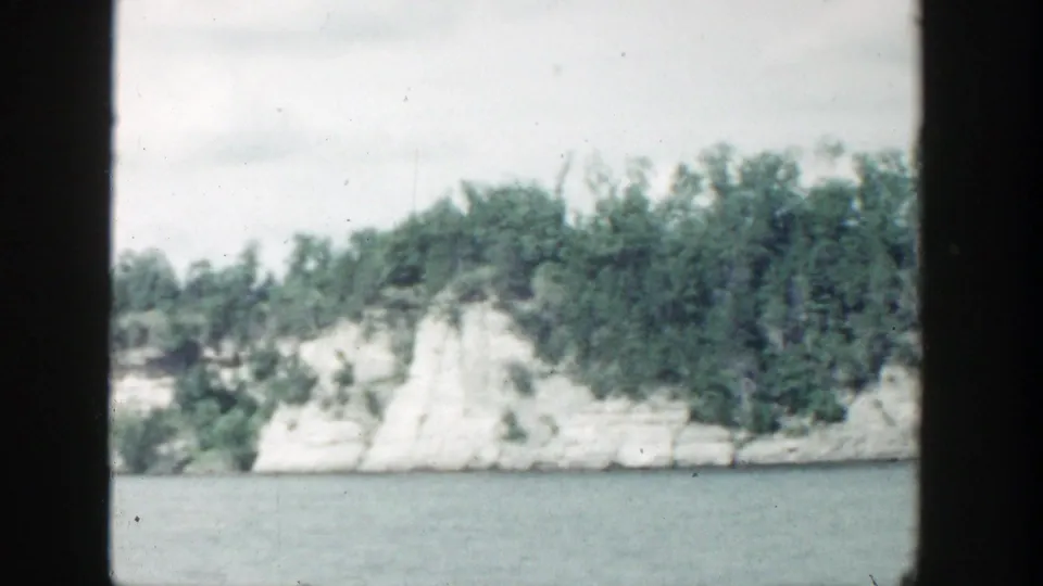 1964 Wisconsin lakeside view with a pale cliff face, green tree cover, calm water, and visible 8mm frame borders.
