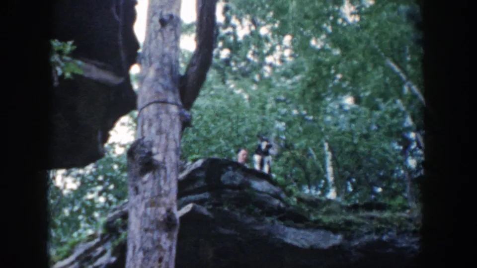 1964 Wisconsin home-movie frame with two distant people on a rocky wooded outcrop beside the lakeside setting.