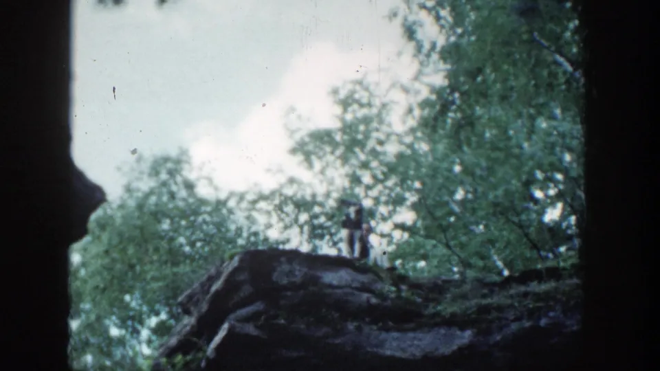 A distant person stands or sits on a rocky forest ridge in Wisconsin, with the dog less distinct in this 8mm frame.