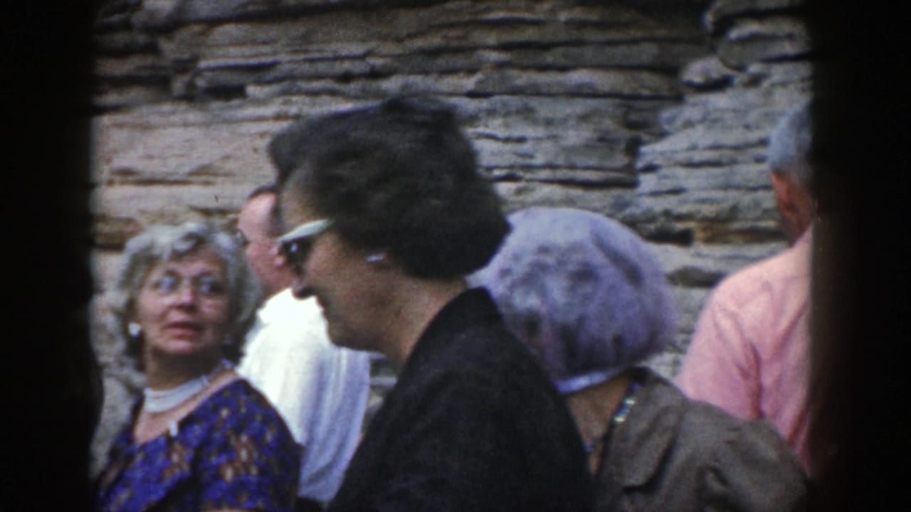 1964 Wisconsin 8mm frame showing adults in 1960s attire gathered beside layered rock, with faces visible in a crowded visitor scene.