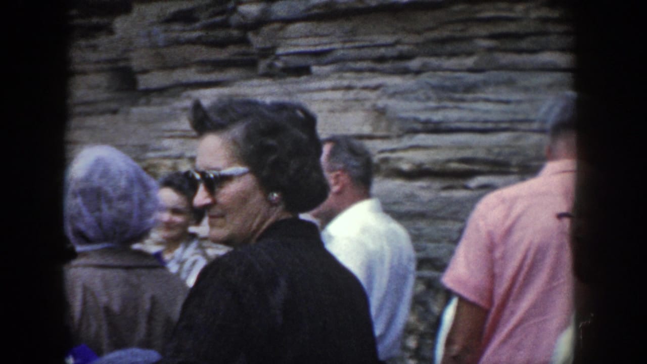 1964 Wisconsin home-movie frame of adults near a layered rock wall, including a woman in sunglasses and black outerwear looking toward the group.