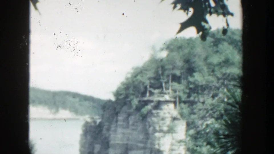 1964 Wisconsin 8mm landscape view showing a wooded rock outcrop above calm lake water, with leaves and film gate borders framing the shot.