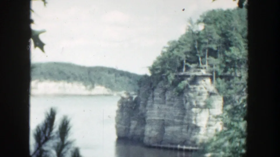 1964 Wisconsin 8mm frame of a tree-covered rock cliff beside calm lake water, with soft focus, faded color, and visible film borders.