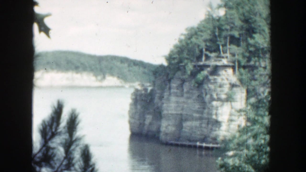 Calm water below a wooded limestone cliff, seen through dark foreground trees.