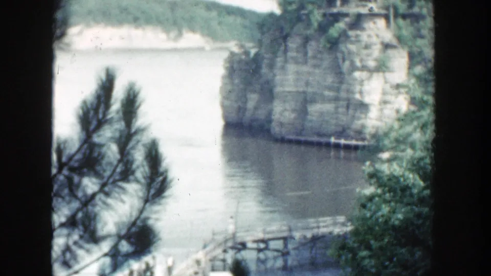 1964 Wisconsin 8mm frame with a small wooden bridge or dock at the lower edge beneath calm water and a tree-covered limestone cliff.