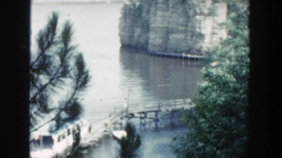 Vintage Wisconsin 8mm view of a small wooden bridge or dock near still water, framed by foliage and a limestone cliff.