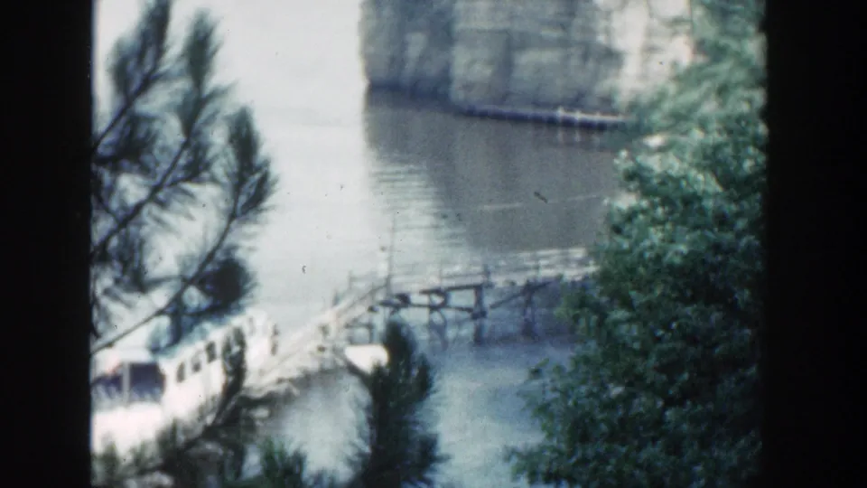1964 Wisconsin 8mm frame of a white lake tour boat by a wooden pier, seen through pine branches and a dark window-like frame.